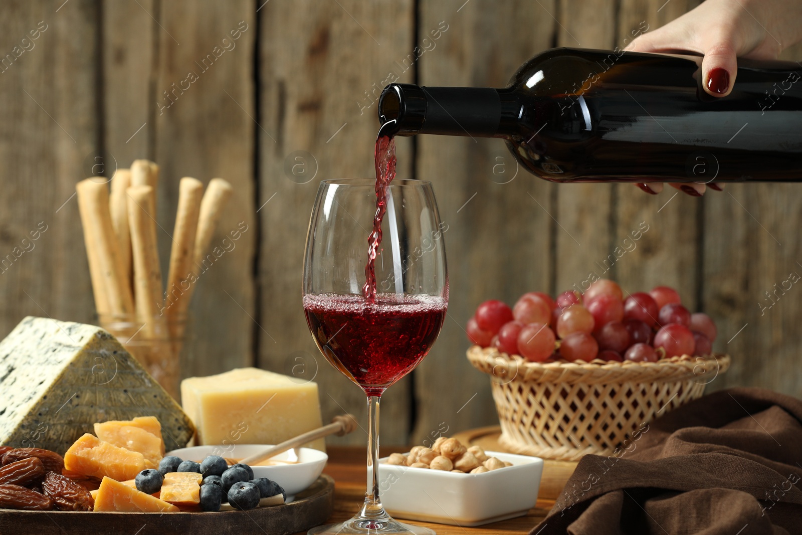 Photo of Woman pouring red wine into glass at table with different snacks, closeup