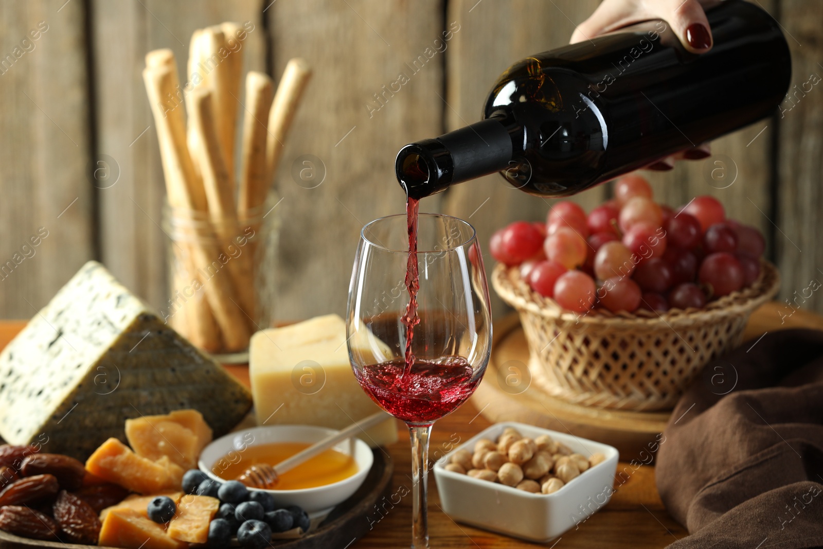 Photo of Woman pouring red wine into glass at table with different snacks, closeup