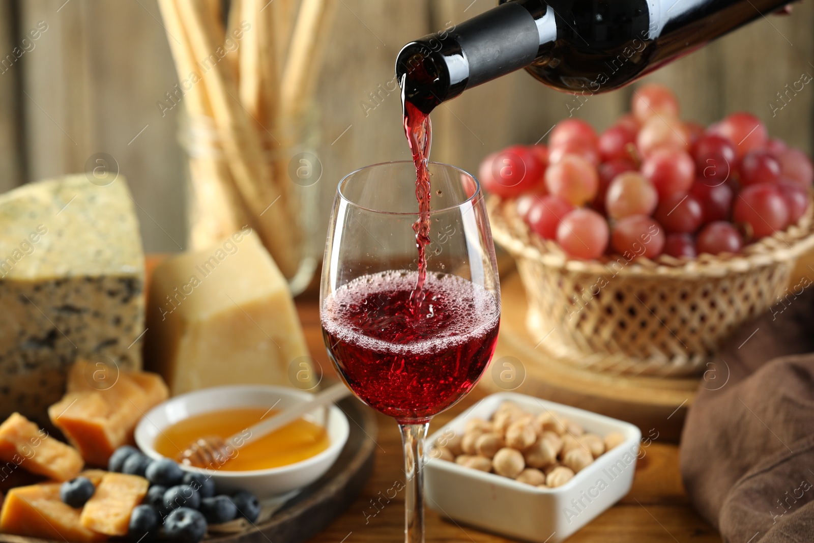 Photo of Pouring red wine into glass at table with different snacks, closeup