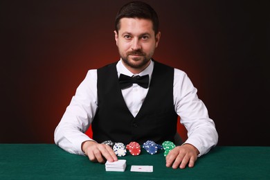 Photo of Professional croupier at gambling table with playing cards and casino chips against dark red background