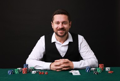 Photo of Professional croupier at gambling table with playing cards, casino chips and dice against black background