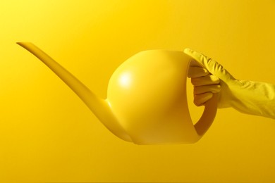 Photo of Woman in rubber glove holding plastic watering can on yellow background, closeup