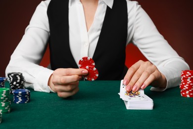 Photo of Professional croupier with casino chips and playing cards at gambling table on color background, closeup