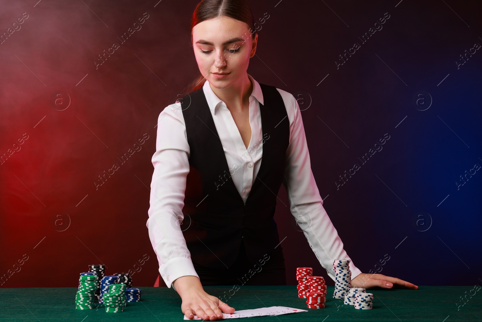 Photo of Professional croupier with casino chips and playing cards at gambling table on color background with smoke