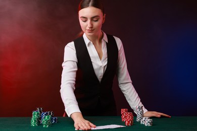 Photo of Professional croupier with casino chips and playing cards at gambling table on color background with smoke