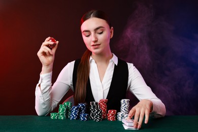 Professional croupier with casino chips and playing cards at gambling table on color background with smoke