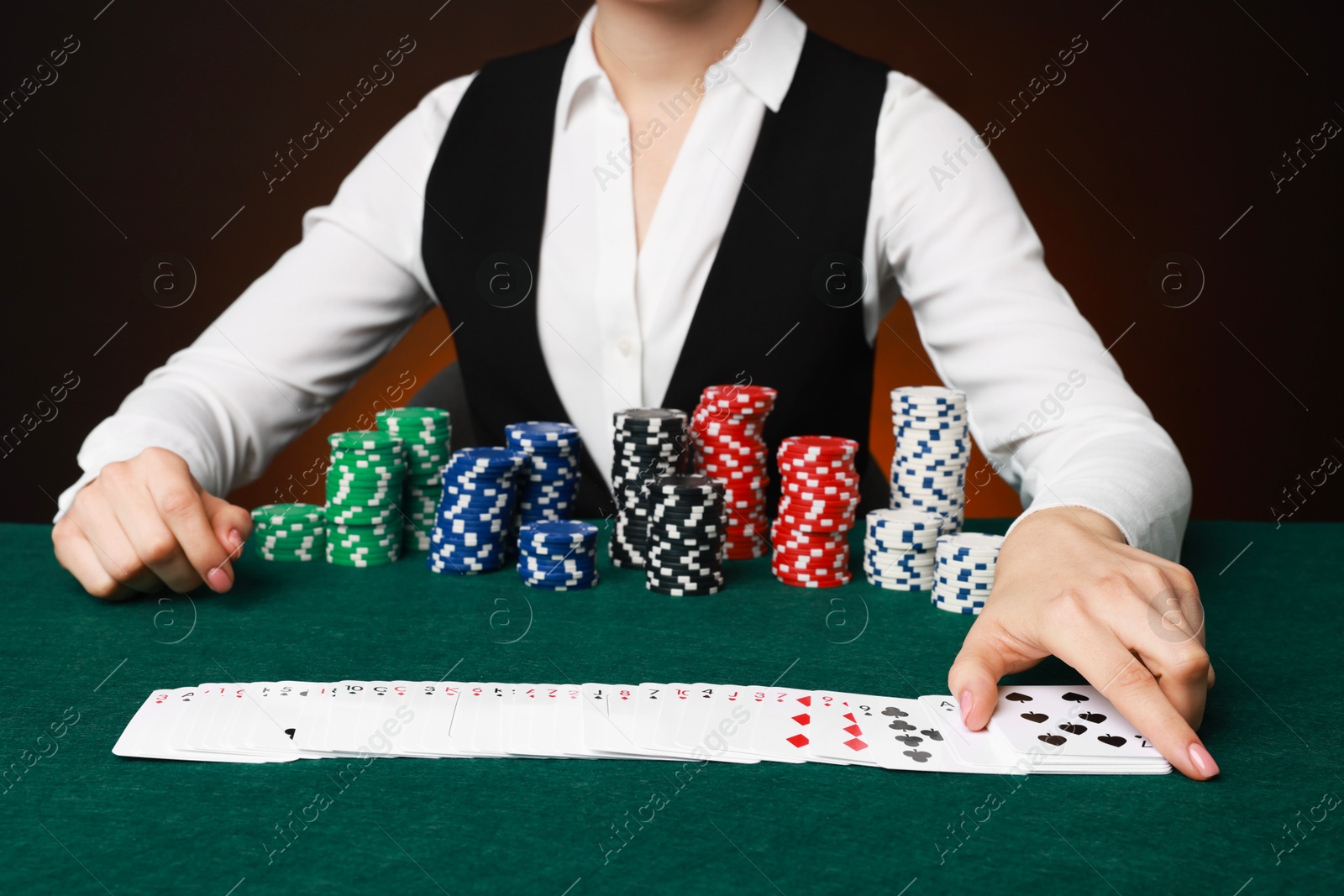 Photo of Professional croupier with casino chips and playing cards at gambling table on color background, closeup