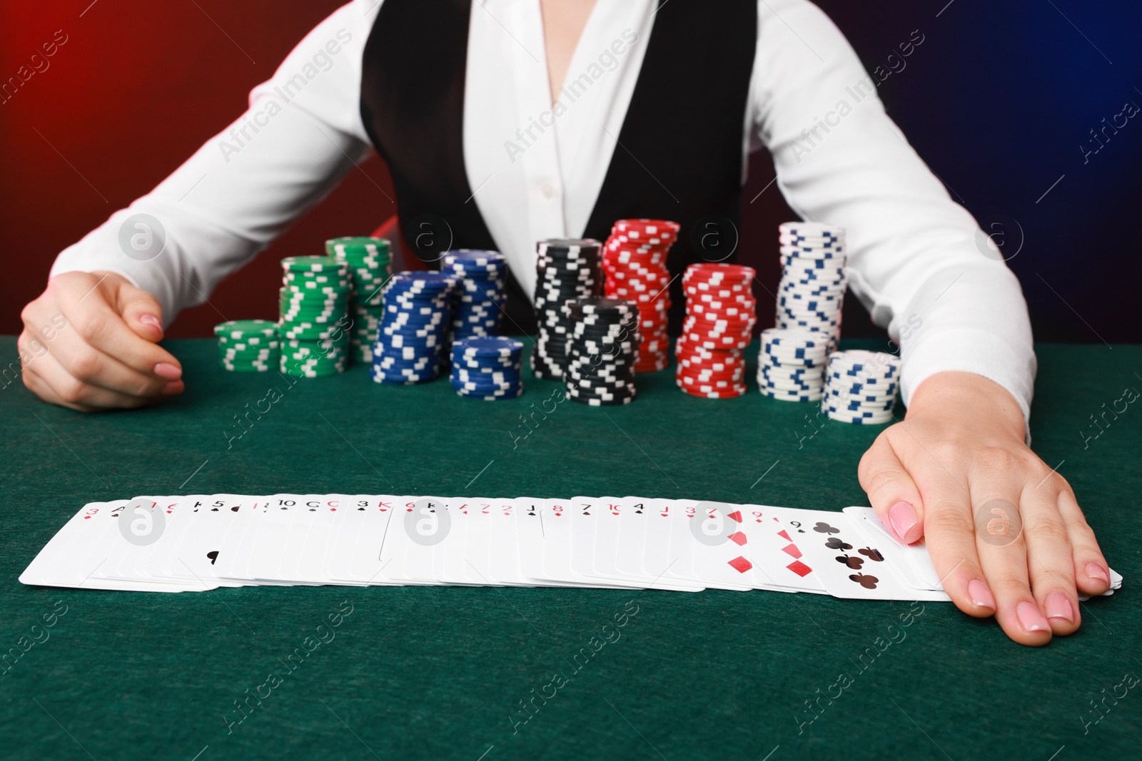 Photo of Professional croupier with casino chips and playing cards at gambling table on color background, closeup