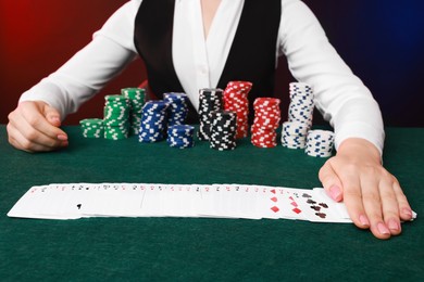 Professional croupier with casino chips and playing cards at gambling table on color background, closeup