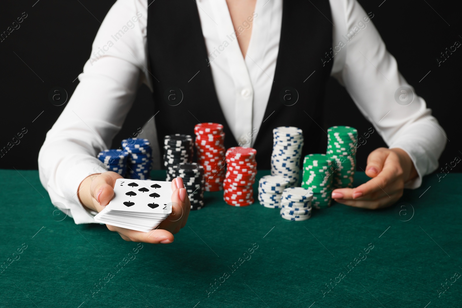 Photo of Professional croupier with casino chips and playing cards at gambling table on black background, closeup