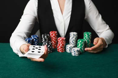 Professional croupier with casino chips and playing cards at gambling table on black background, closeup