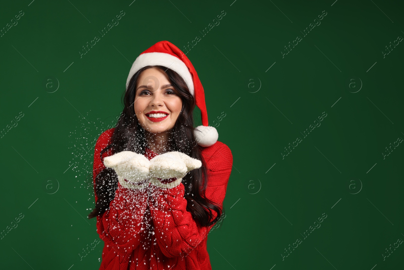 Photo of Attractive woman in Santa hat blowing snow off hands on green background, space for text. Christmas celebration