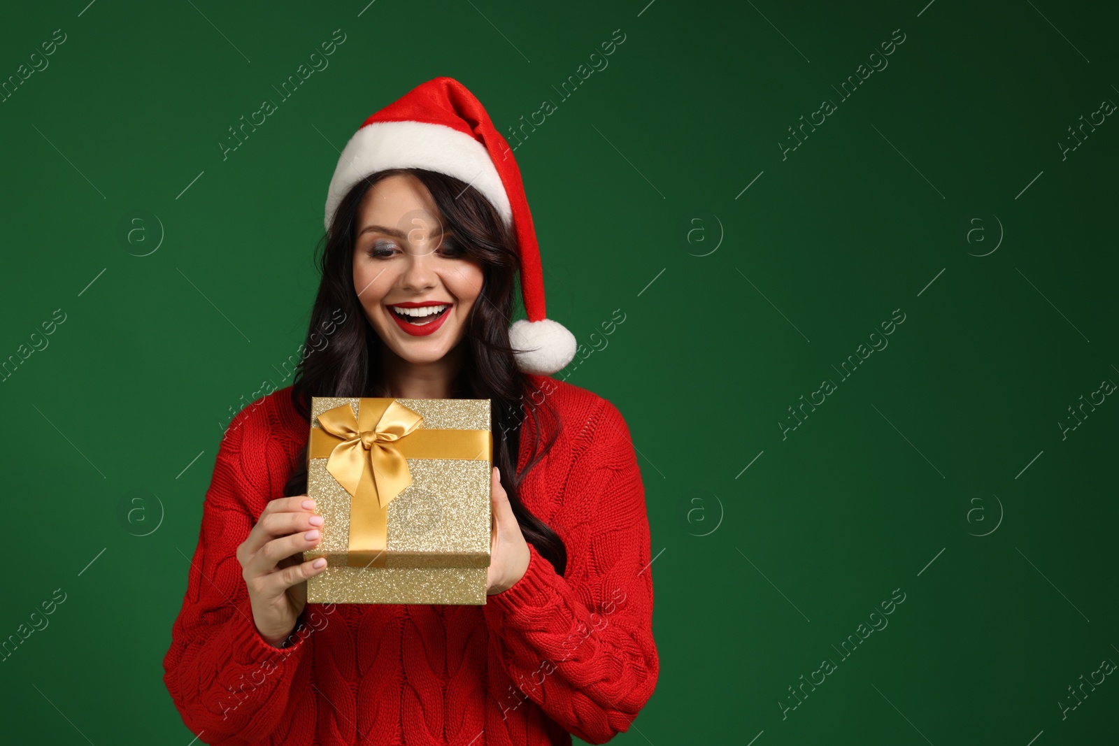 Photo of Attractive woman in Santa hat opening Christmas gift on green background, space for text