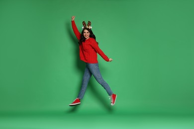 Photo of Happy woman in sweater and reindeer headband jumping on green background. Christmas celebration