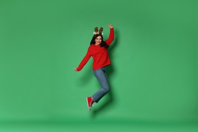 Photo of Happy woman in sweater and reindeer headband jumping on green background. Christmas celebration