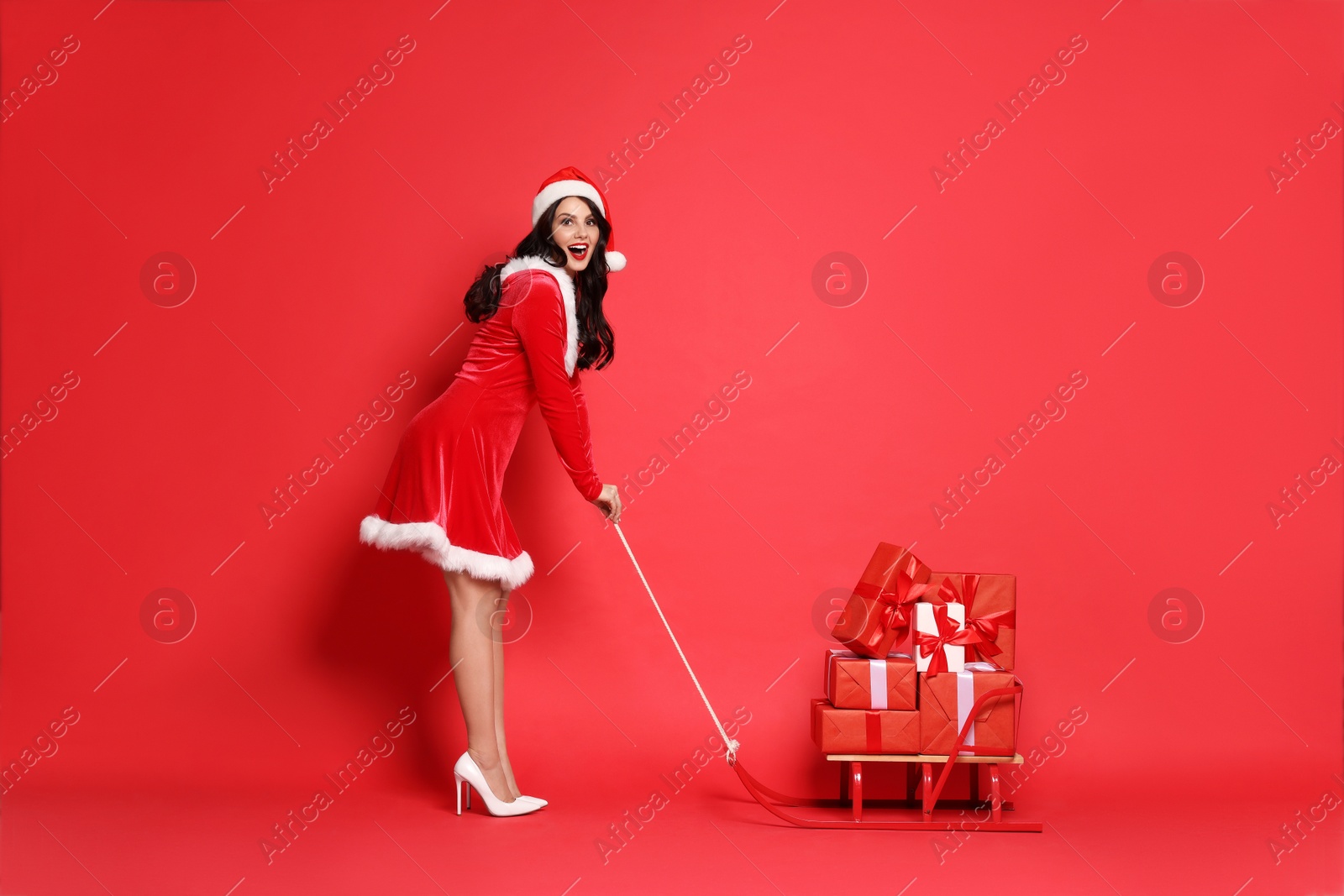 Photo of Beautiful woman in Mrs Claus costume with Christmas gifts on sledge against red background