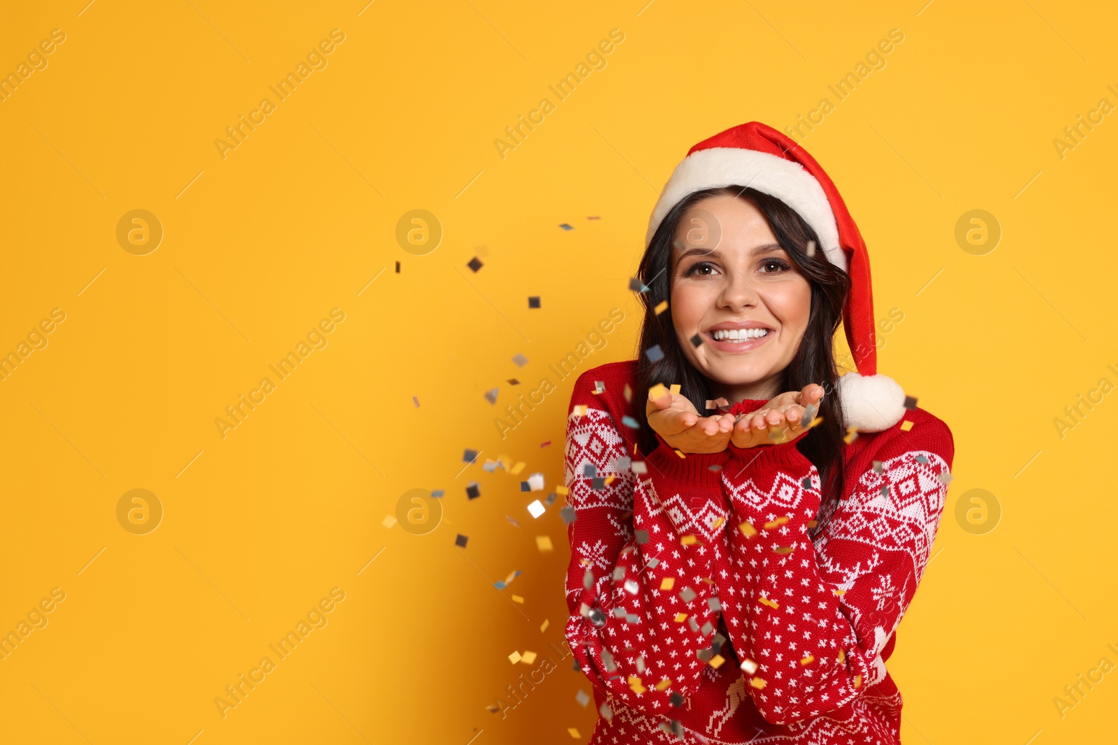 Photo of Beautiful woman in Santa hat and Christmas sweater with confetti on yellow background, space for text