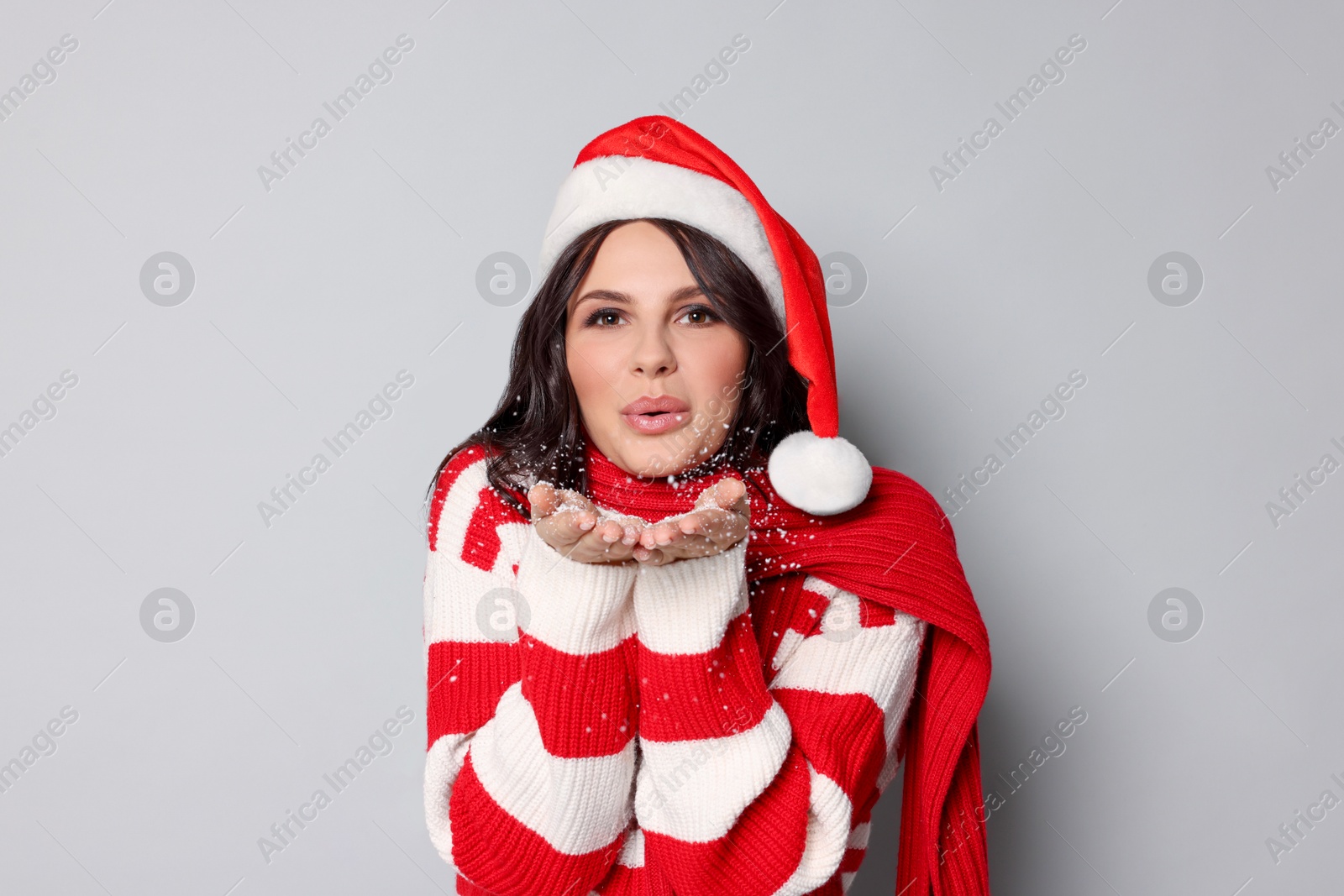 Photo of Attractive woman in Santa hat blowing snow off hands on light grey background. Christmas celebration