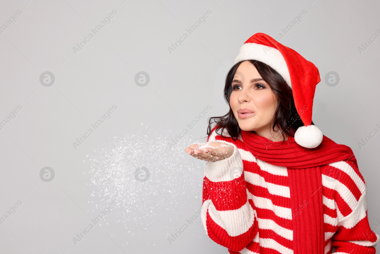 Photo of Attractive woman in Santa hat blowing snow off hands on light grey background, space for text. Christmas celebration