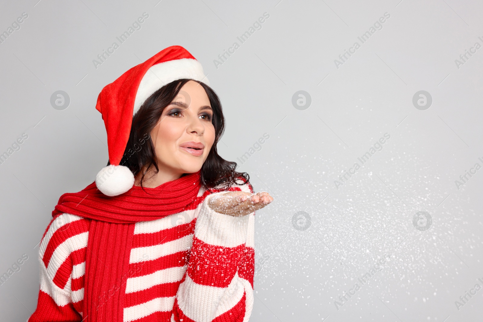 Photo of Attractive woman in Santa hat blowing snow off hands on light grey background, space for text. Christmas celebration