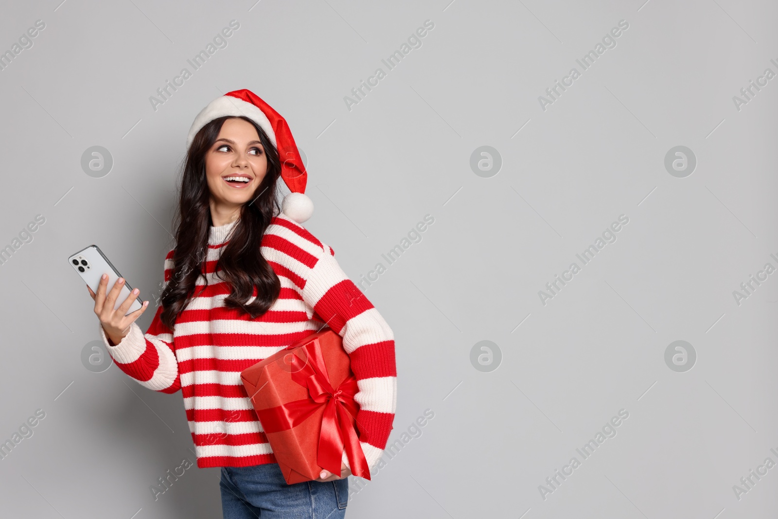 Photo of Woman in Santa hat with Christmas gift and smartphone on light grey background, space for text