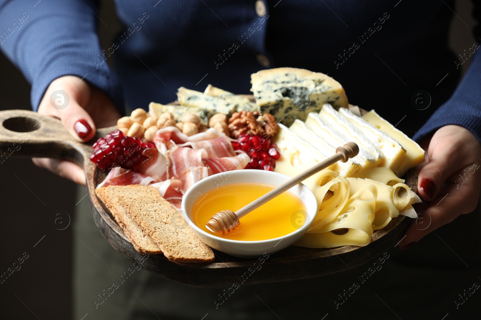 Photo of Woman holding board with different types of delicious cheese and other snacks on gray background, closeup