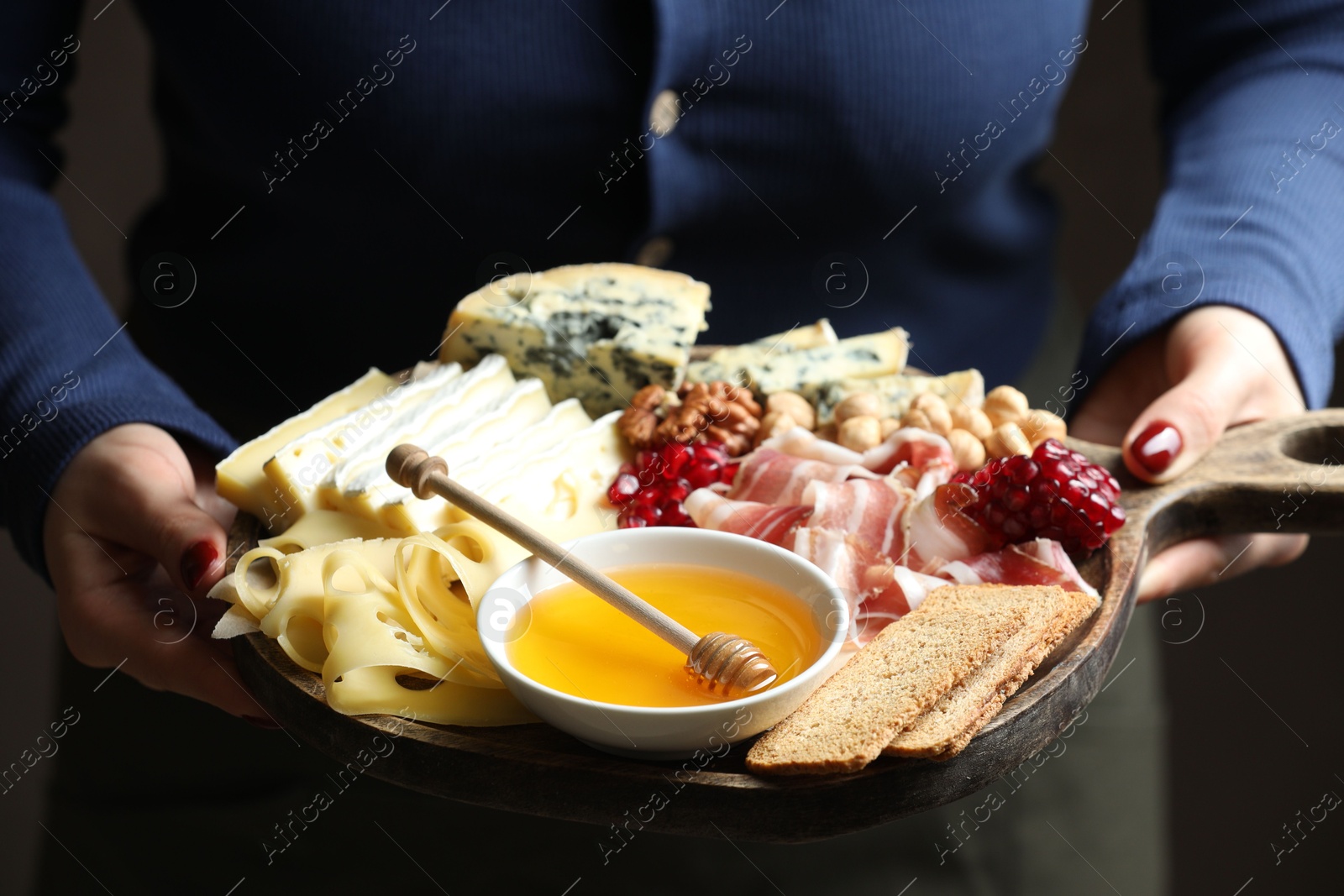 Photo of Woman holding board with different types of delicious cheese and other snacks on gray background, closeup