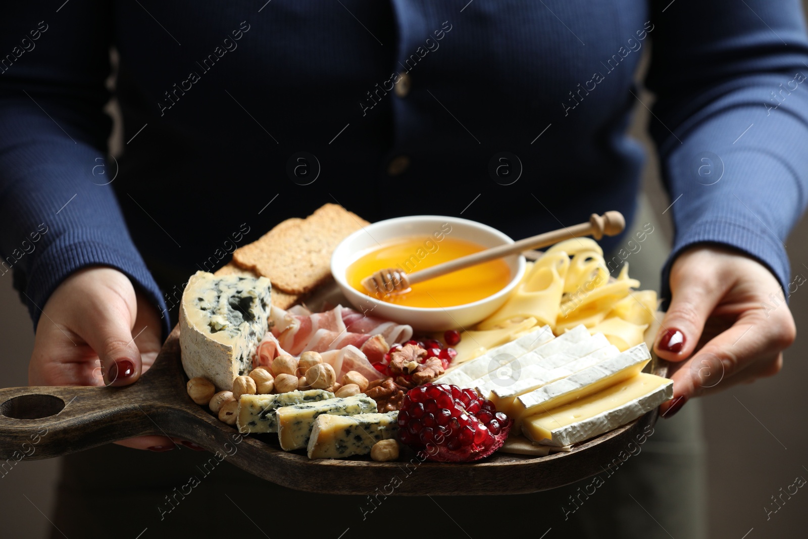 Photo of Woman holding board with different types of delicious cheese and other snacks on gray background, closeup