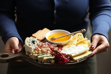 Photo of Woman holding board with different types of delicious cheese and other snacks on gray background, closeup