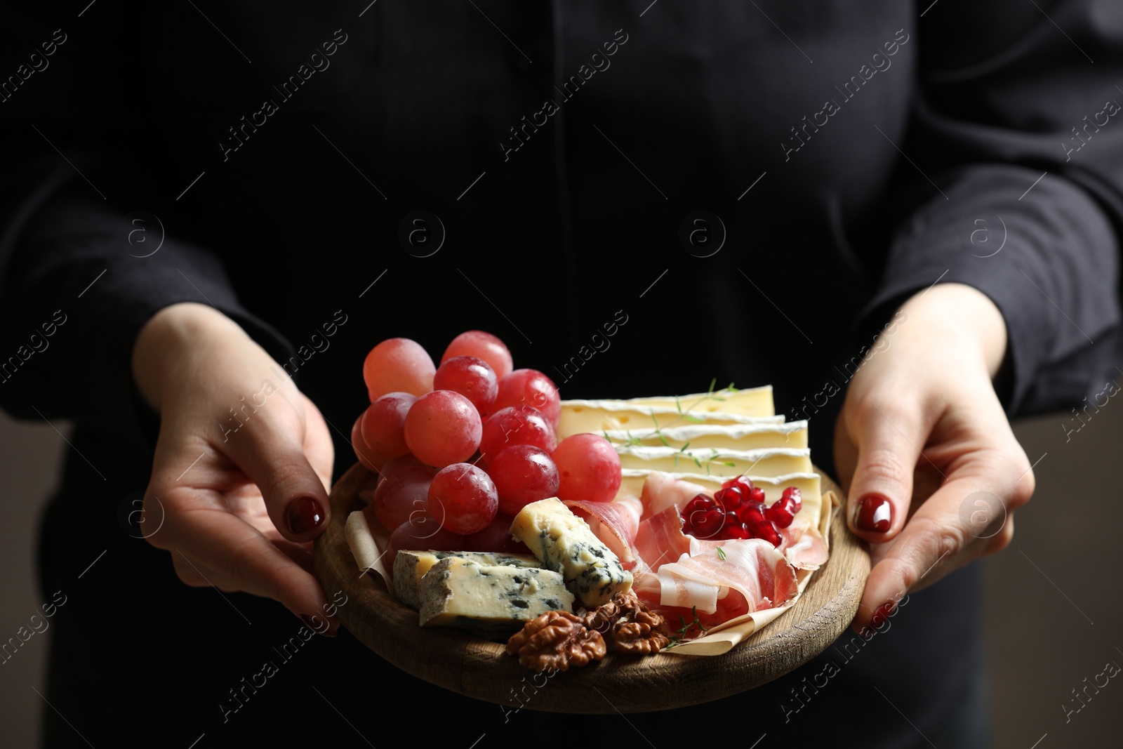 Photo of Woman holding board with different types of delicious cheese and other snacks on gray background, closeup