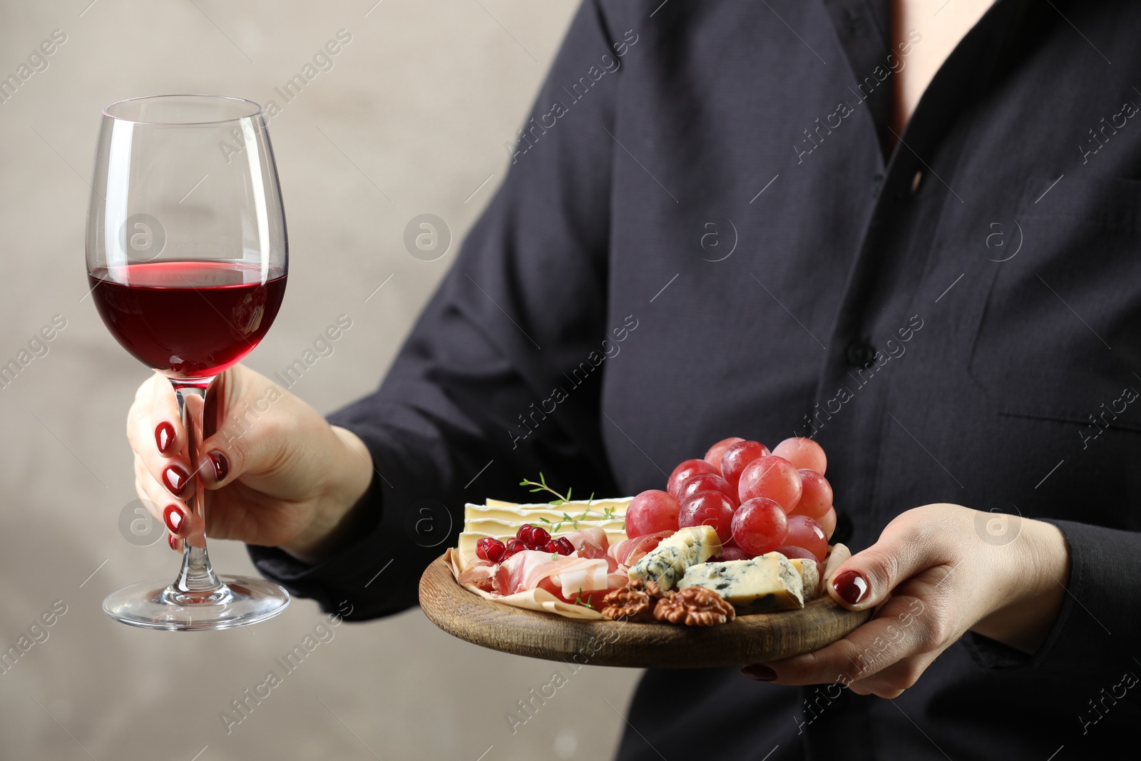 Photo of Woman holding board with different types of delicious cheese, other snacks and wine on gray background, closeup