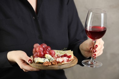 Photo of Woman holding board with different types of delicious cheese, other snacks and wine on gray background, closeup