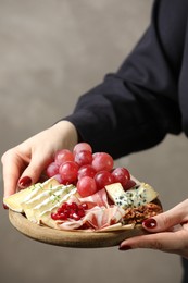 Photo of Woman holding board with different types of delicious cheese and other snacks on gray background, closeup