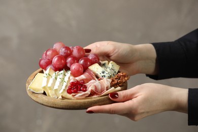 Photo of Woman holding board with different types of delicious cheese and other snacks on gray background, closeup