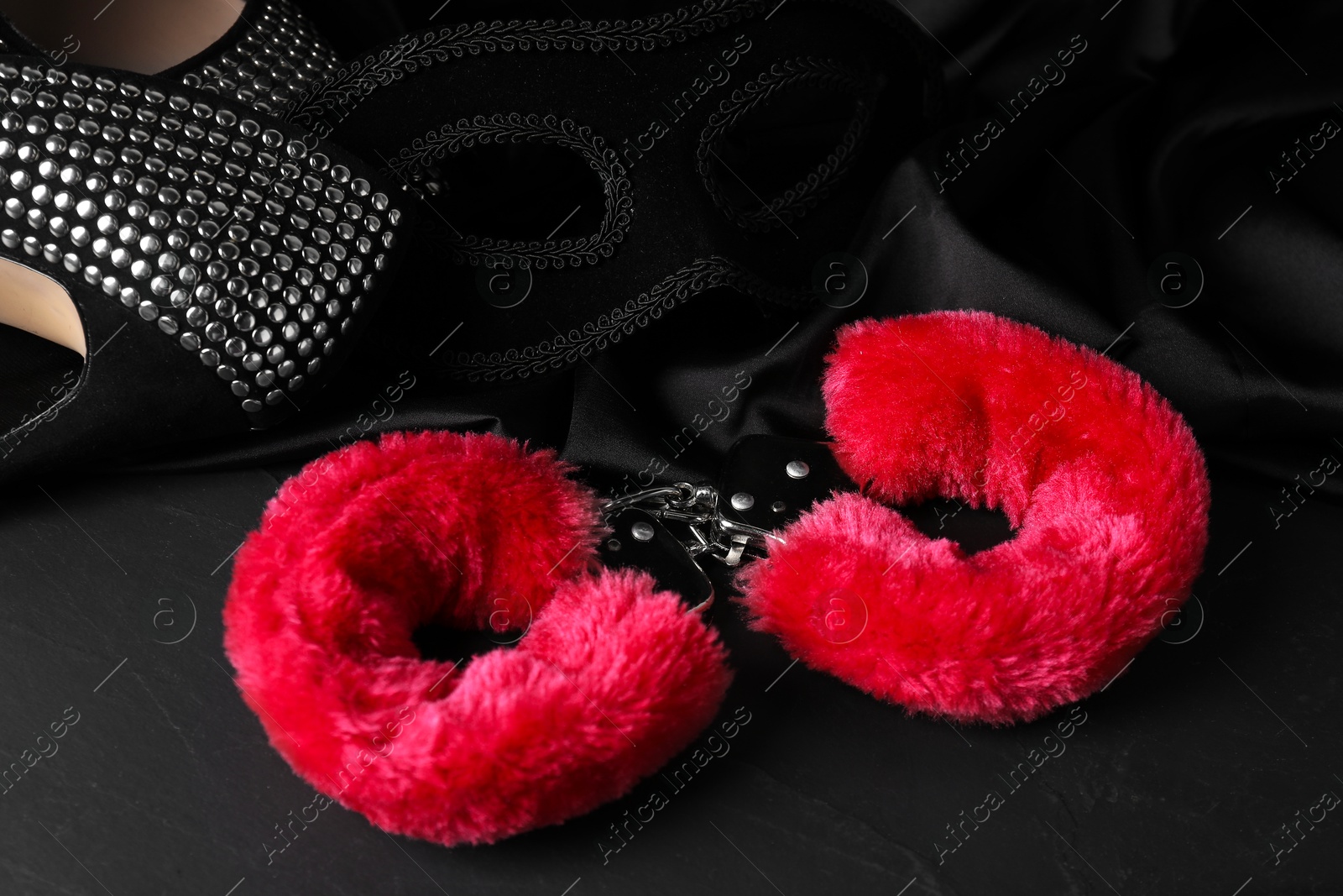 Photo of Red fluffy handcuffs, mask, shoes and fabric on black surface