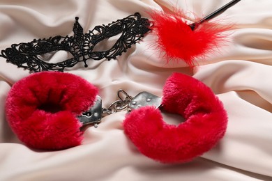 Photo of Red fluffy handcuffs, lace mask and feather on beige fabric, closeup