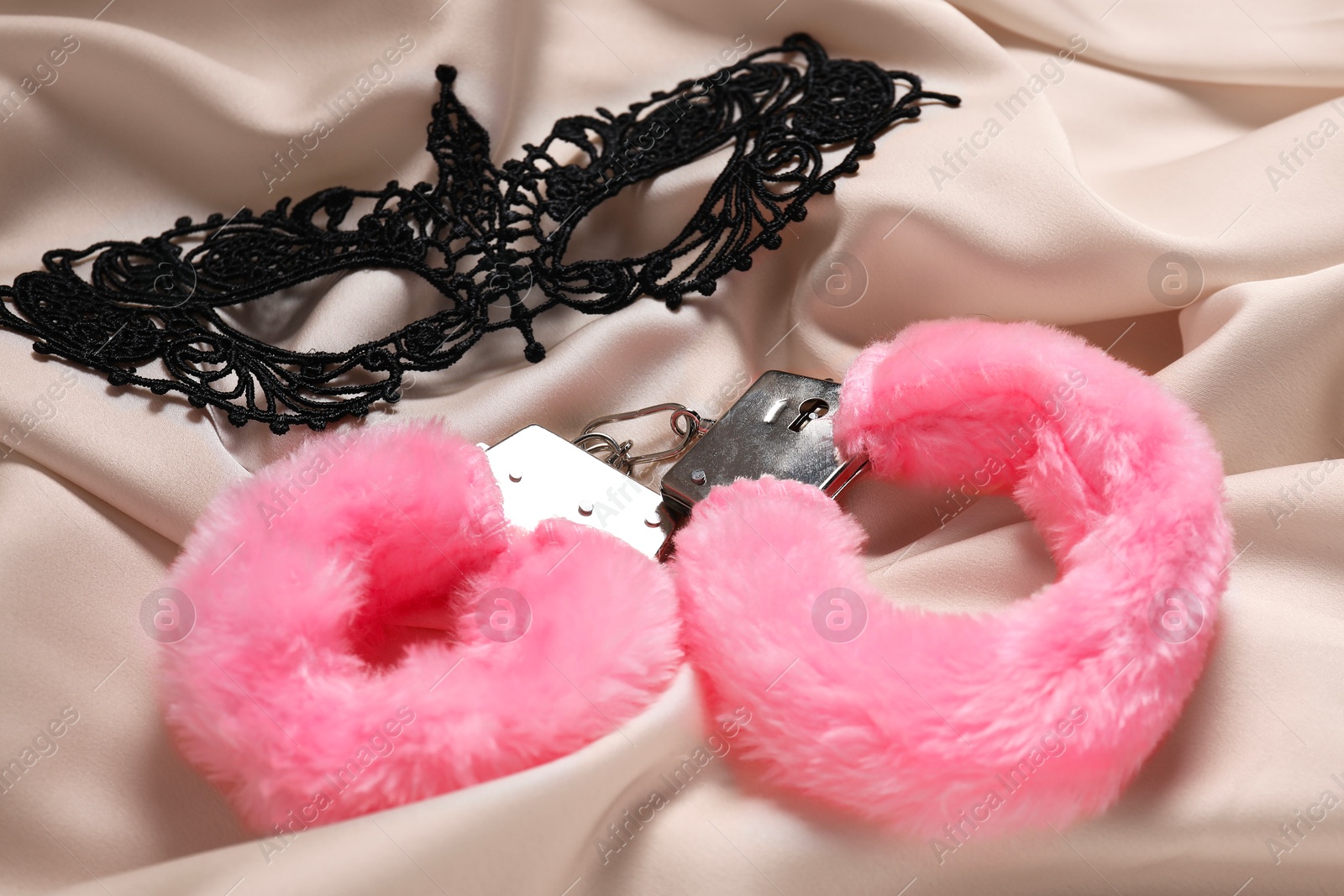 Photo of Pink fluffy handcuffs and lace mask on beige fabric, closeup