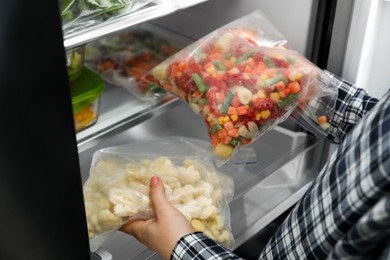 Photo of Woman taking plastic bags with mix of frozen vegetables and cauliflower, from refrigerator, closeup