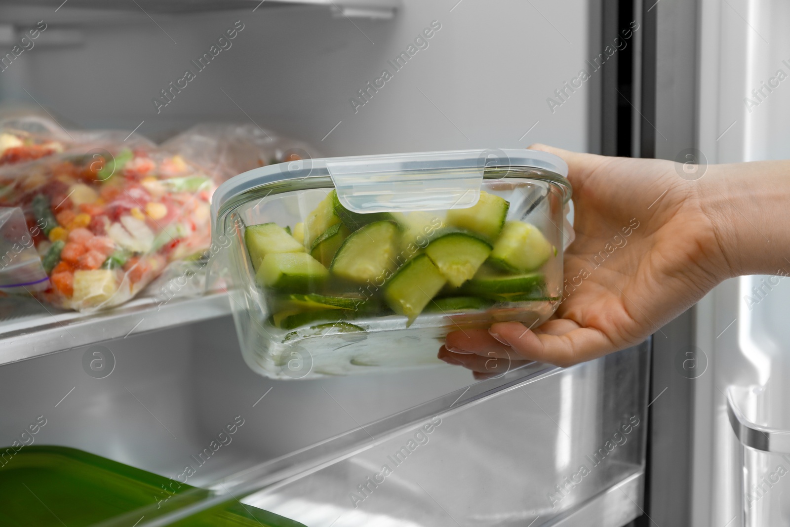 Photo of Woman taking glass container with frozen chopped zucchini from refrigerator, closeup