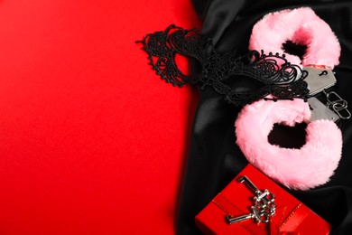 Photo of Pink fluffy handcuffs, lace mask, keys and black fabric on red background, space for text