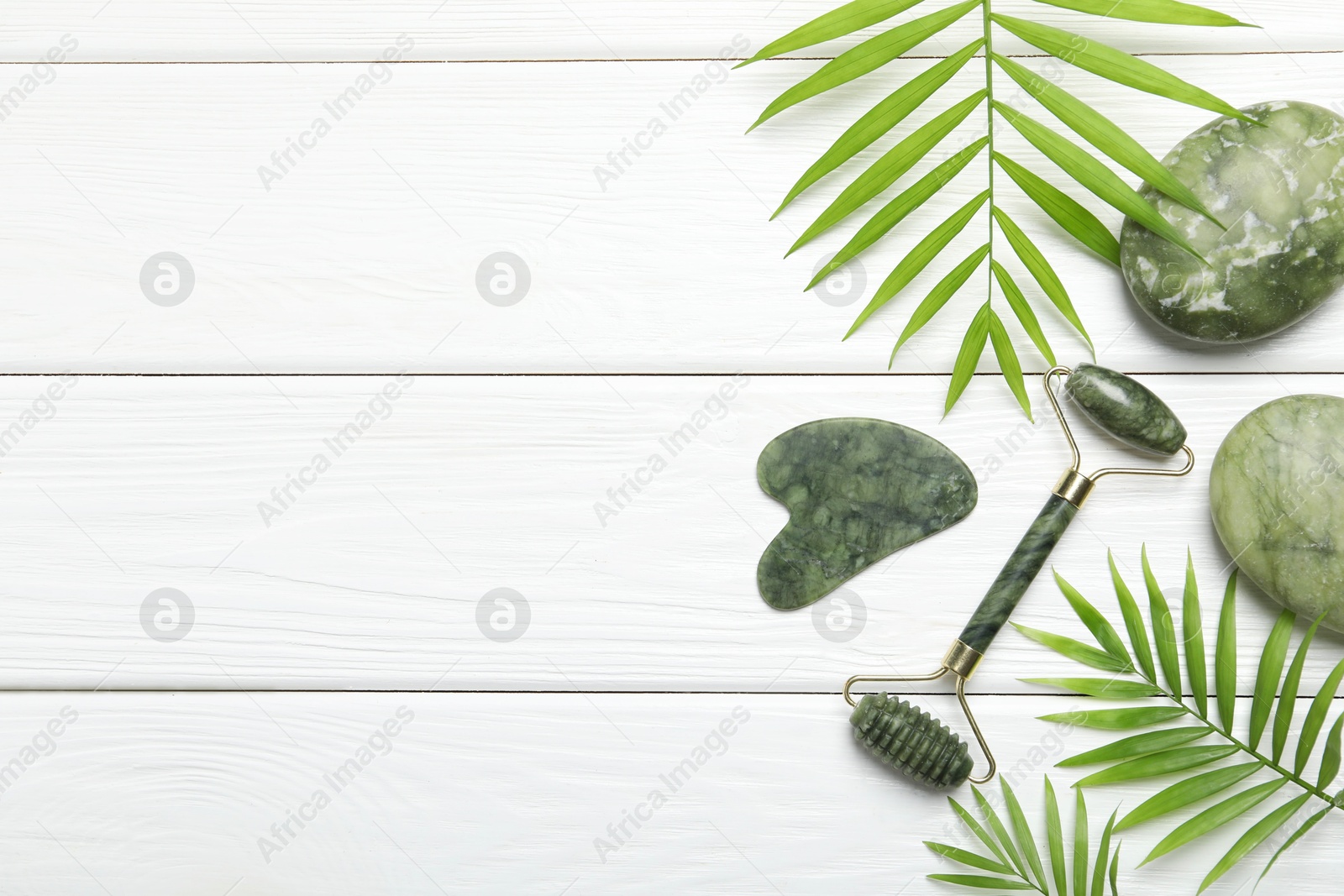 Photo of Face roller, gua sha tool, massage stones and palm leaves on white wooden background, flat lay. Space for text