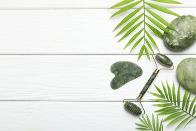 Photo of Face roller, gua sha tool, massage stones and palm leaves on white wooden background, flat lay. Space for text