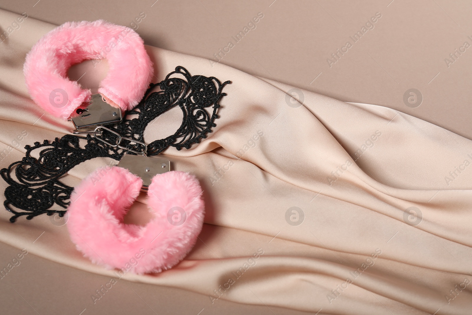 Photo of Pink fluffy handcuffs, lace mask and fabric on dark beige background. Space for text