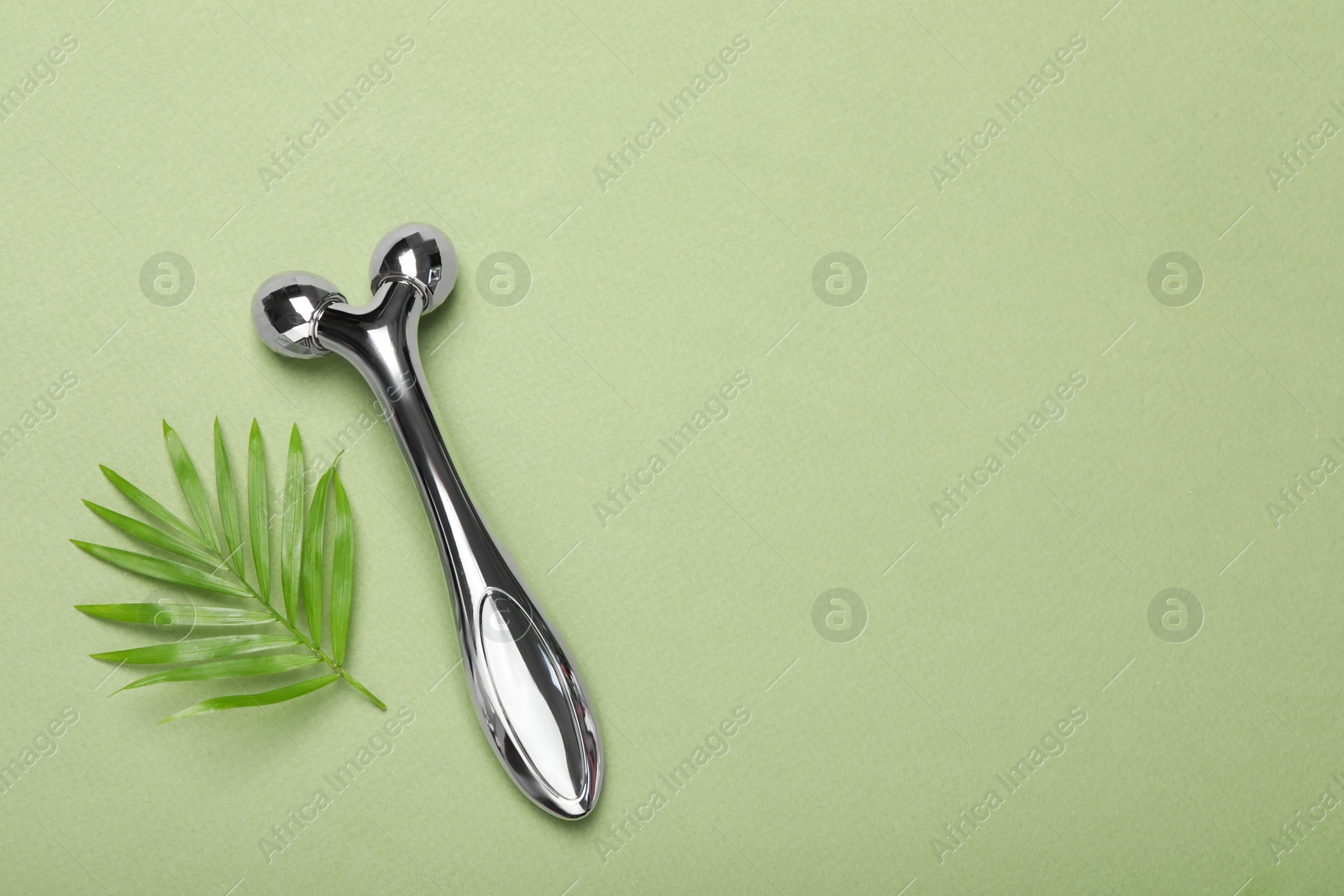 Photo of Metal face roller and palm leaf on green background, flat lay. Space for text