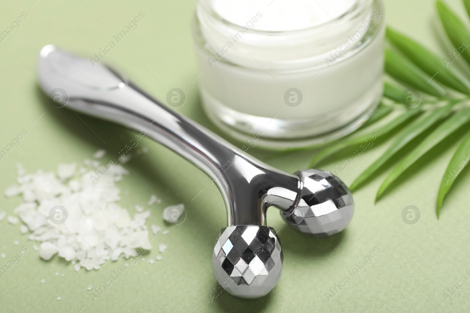 Photo of Metal face roller, cosmetic product, sea salt and palm leaf on green background, closeup