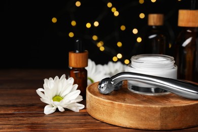 Photo of Metal face roller, cosmetic products and chrysanthemum flower on wooden table against blurred lights, closeup