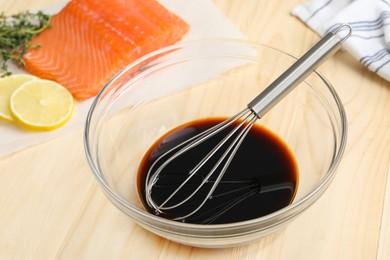 Photo of Soy sauce in bowl, whisk, salmon fillet, thyme and lemon slices on wooden table, closeup