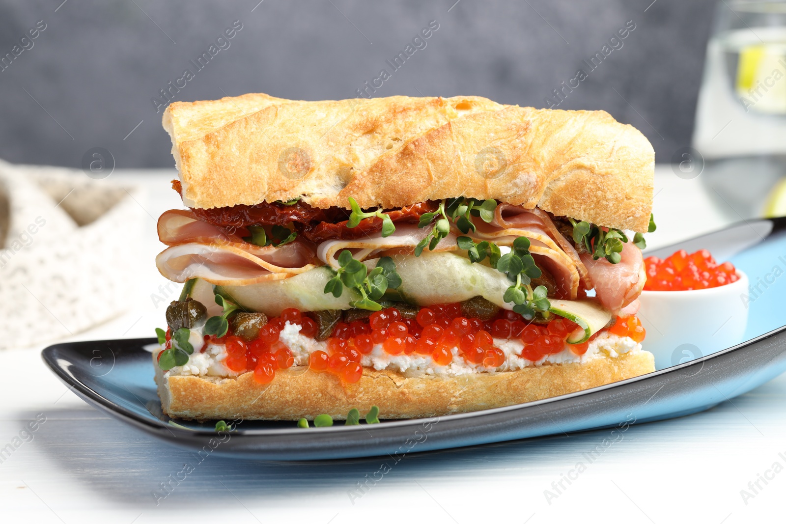 Photo of Delicious baguette sandwich with caviar, ham and capers on white wooden table, closeup