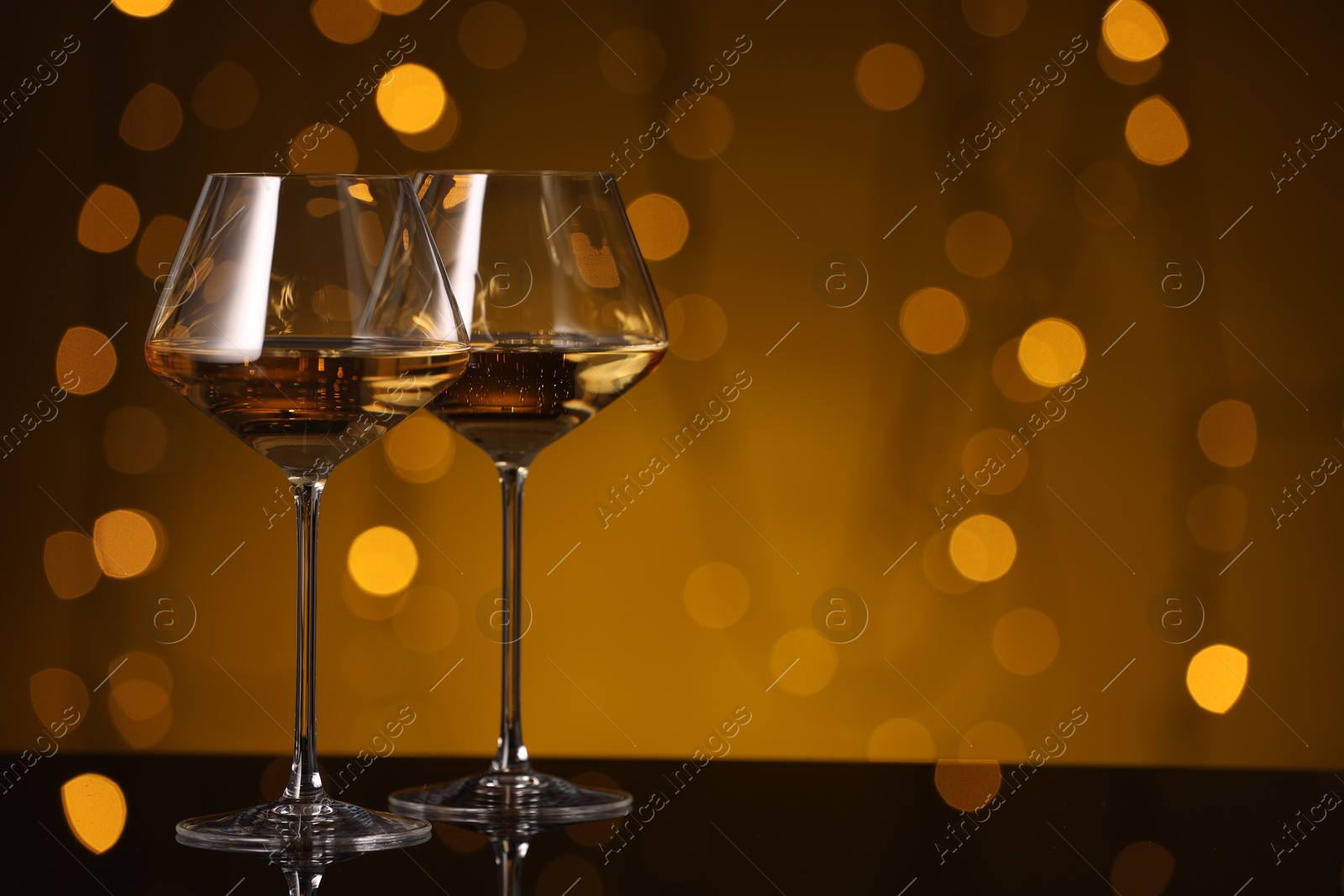 Photo of Tasty white wine in glasses on mirror surface against golden background with blurred lights, space for text. Bokeh effect