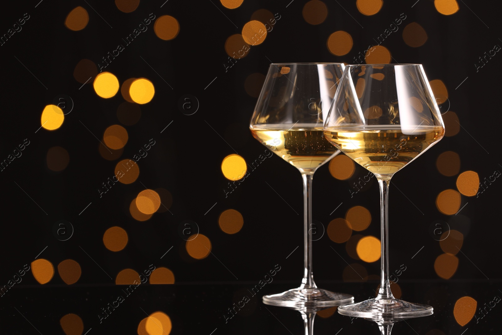 Photo of Tasty white wine in glasses on mirror surface against dark background with blurred lights, space for text. Bokeh effect